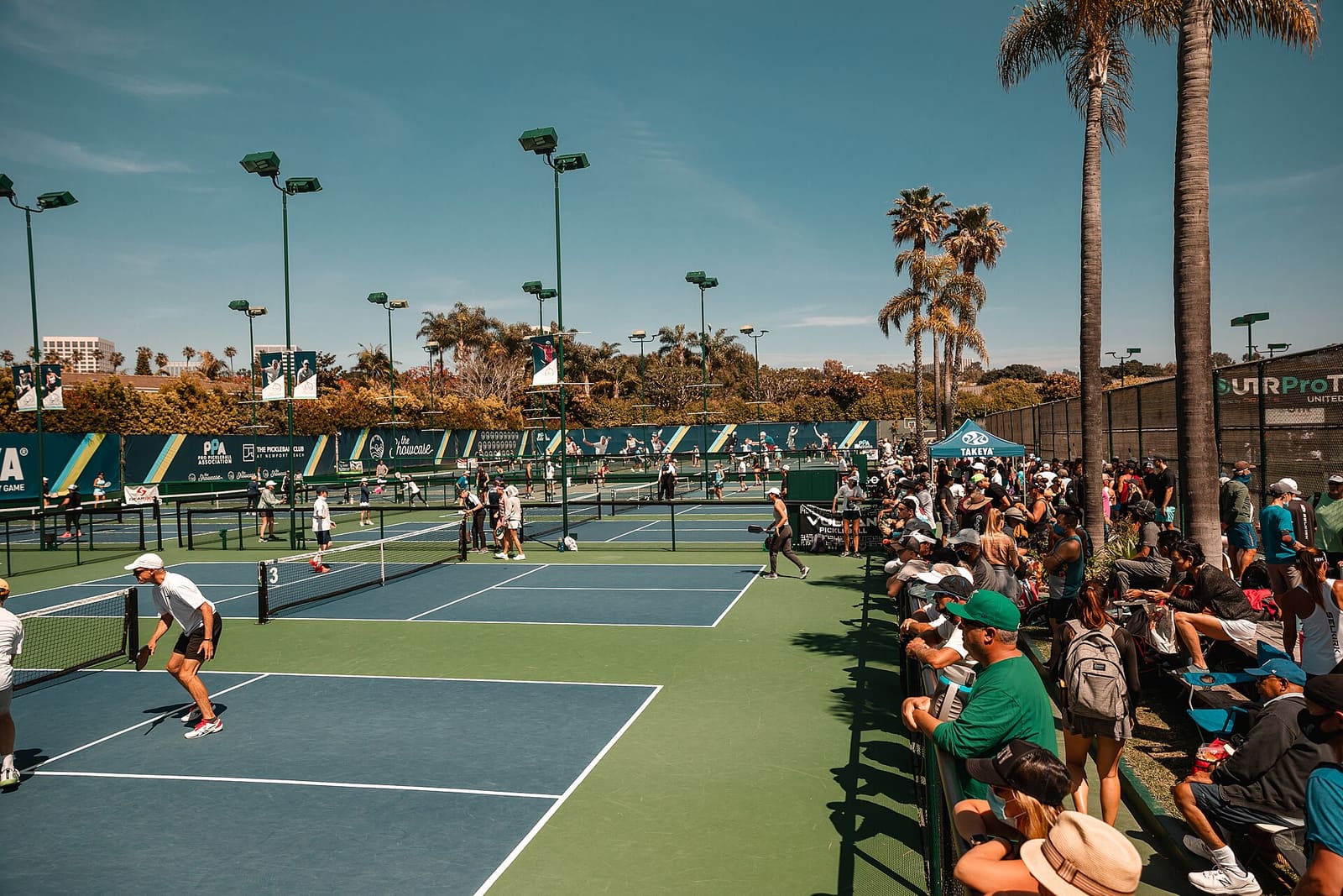 Rows of pickleball courts in play at an outdoor tournament venue, with spectators along the fence.