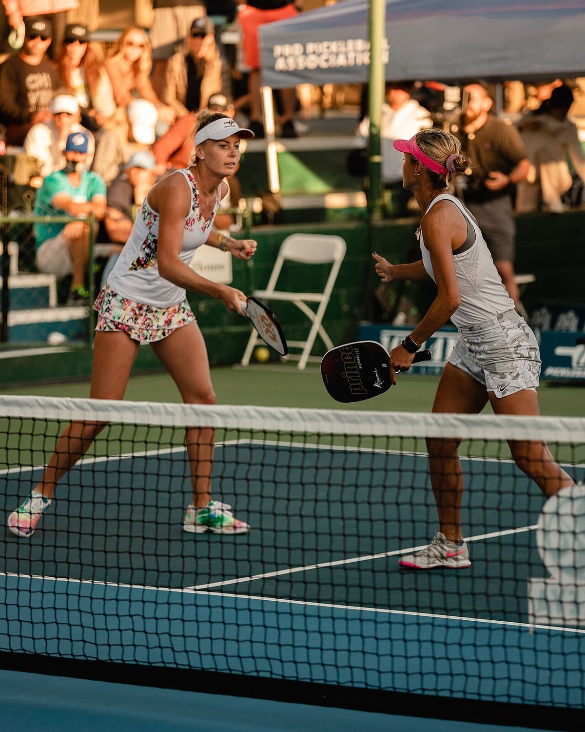 Two doubles players holding the non-volley line at the net, paddles up, during a tournament match.