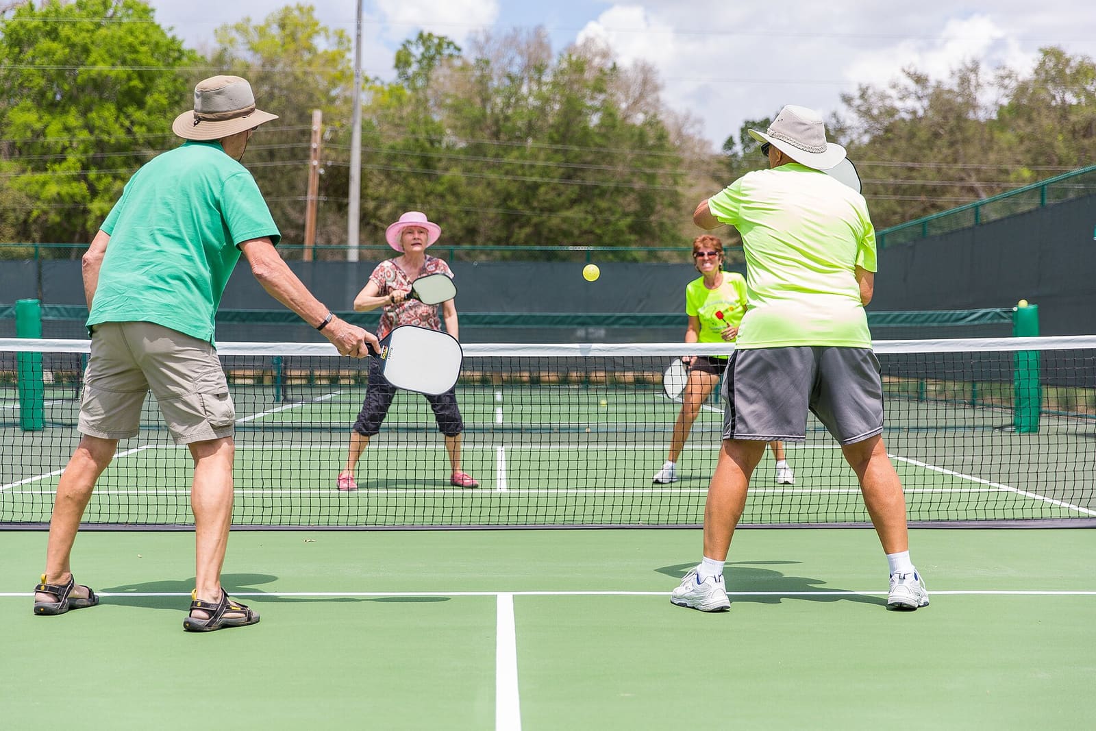 Four players in a doubles game, all standing at the net on an outdoor pickleball court, with the ball in the air between them.